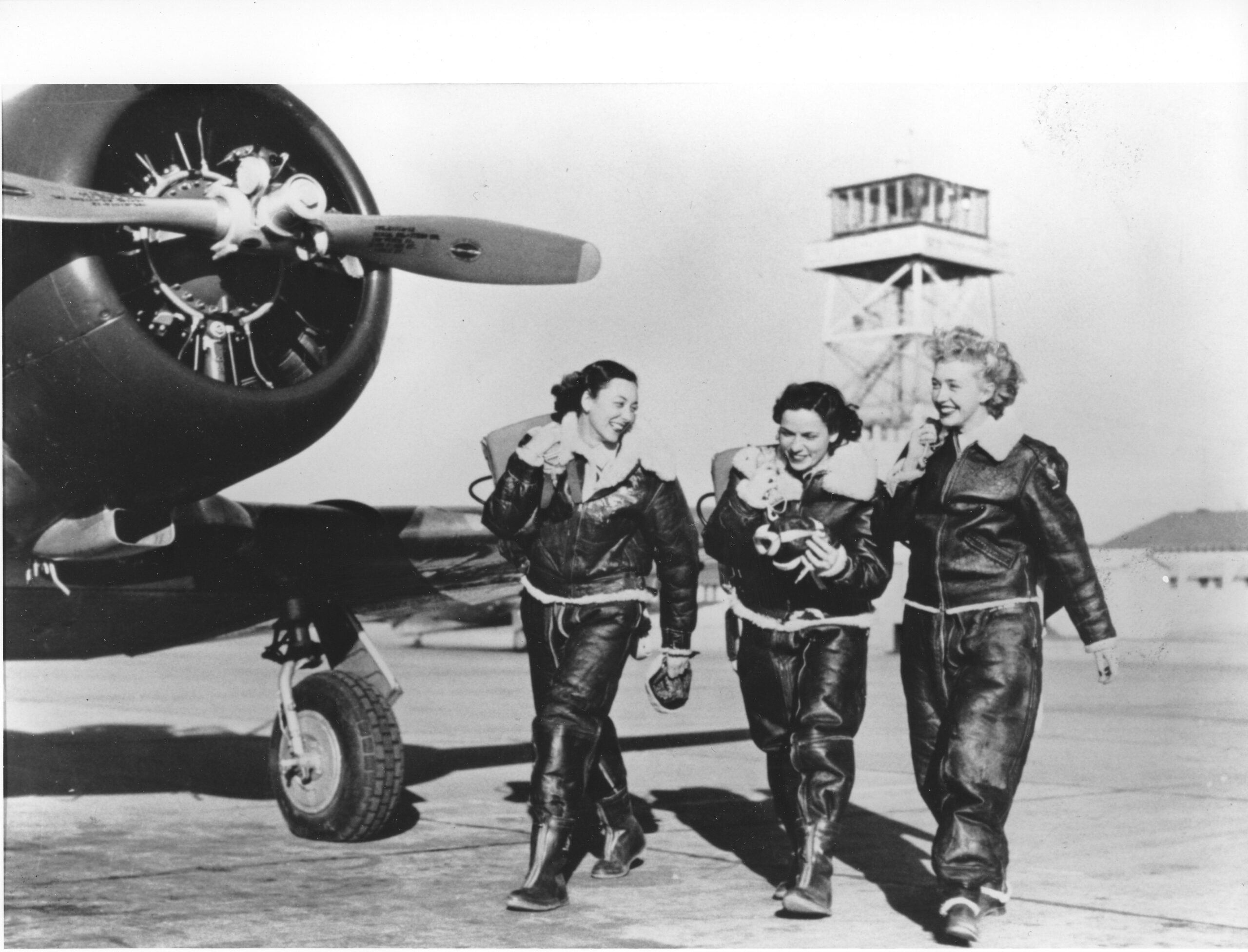 Three women in 1940s airforce attire walk past a plane.