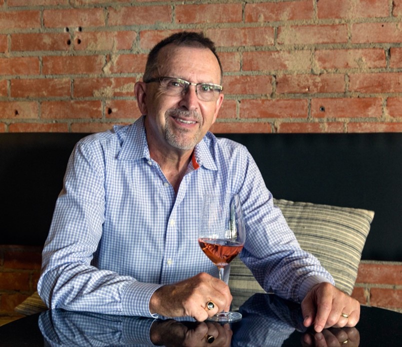 Headshot of Russ Kane. He wears a blue button down shirt and holds a glass of wine. He is smiling.
